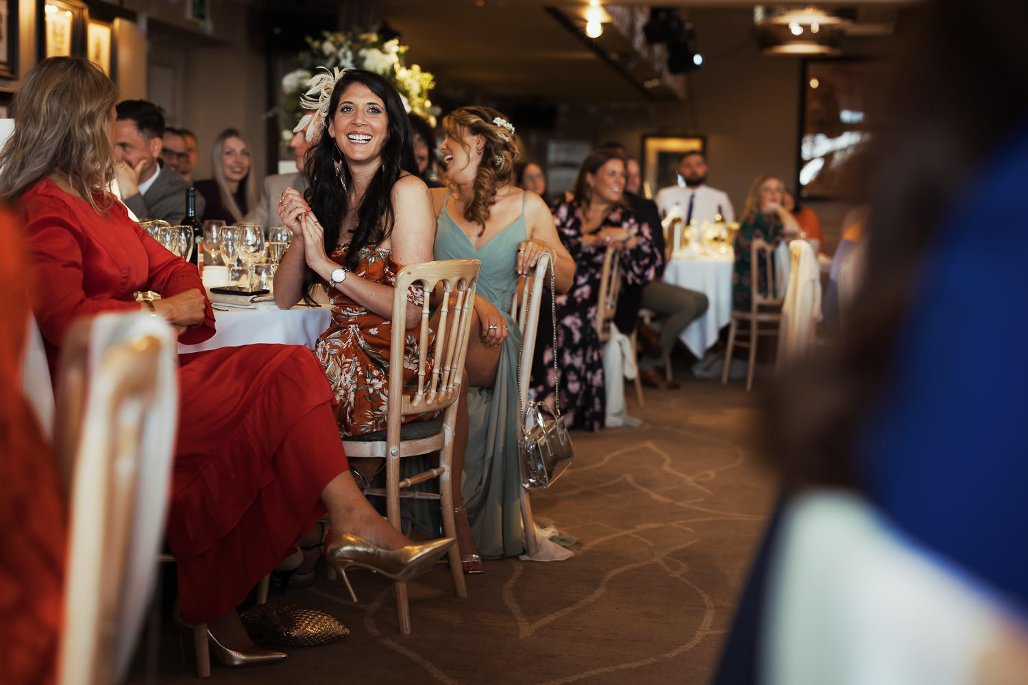 Woman with long dark hair and huge smile, holds her hands together. She's wearing a RUST FLORAL PRINT SATIN DRAPED BARDOT MIDI DRESS from Pretty Little Thing. She's watching the speeches at a wedding reception at The Lion House.