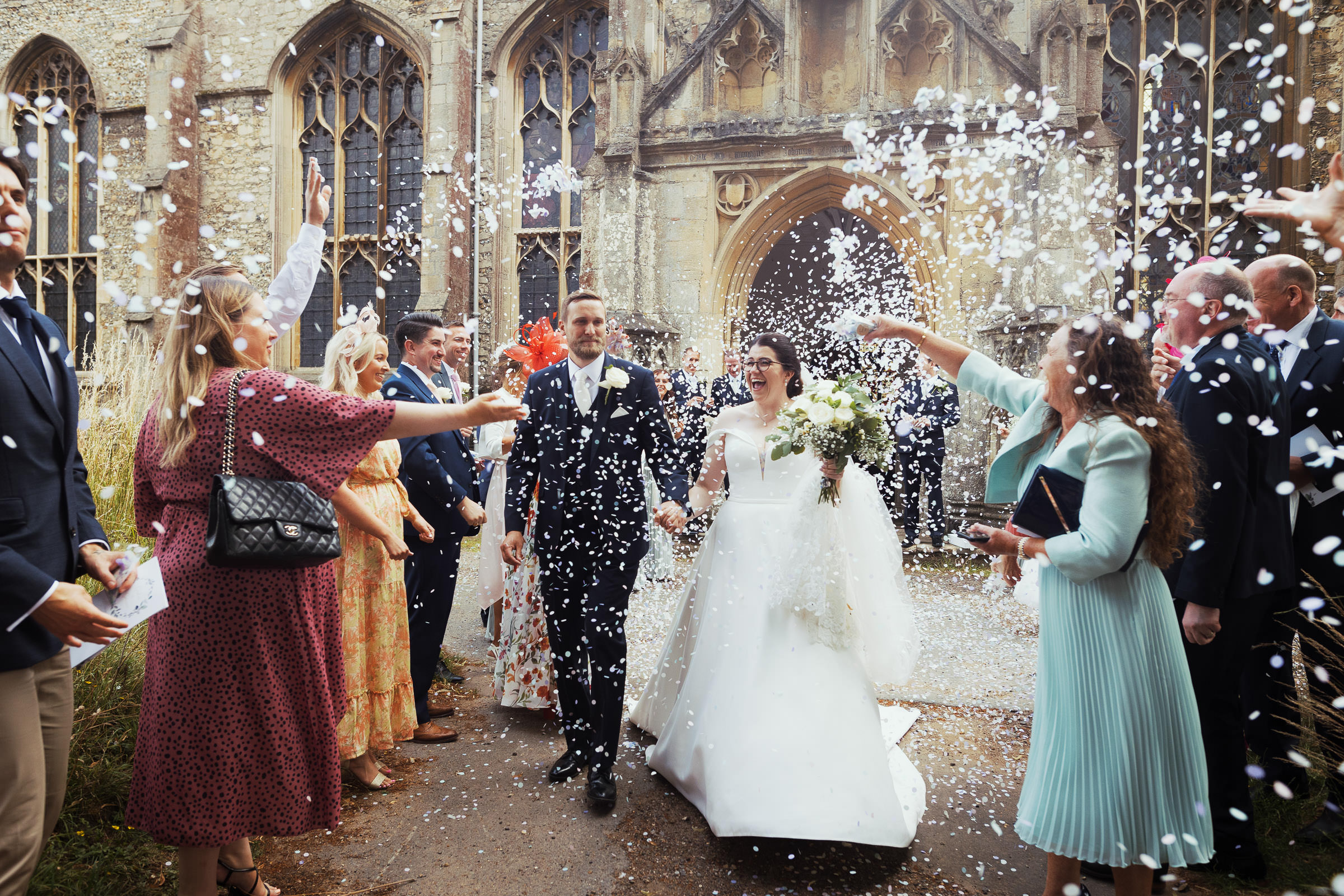 At St Mary's parish church in Bury St Edmund's a new husband and wife walk as guests throw confetti at them. Bride is wearing  Essense of Australia ballgown style wedding dress D2761 from House of Snow bridal shop.