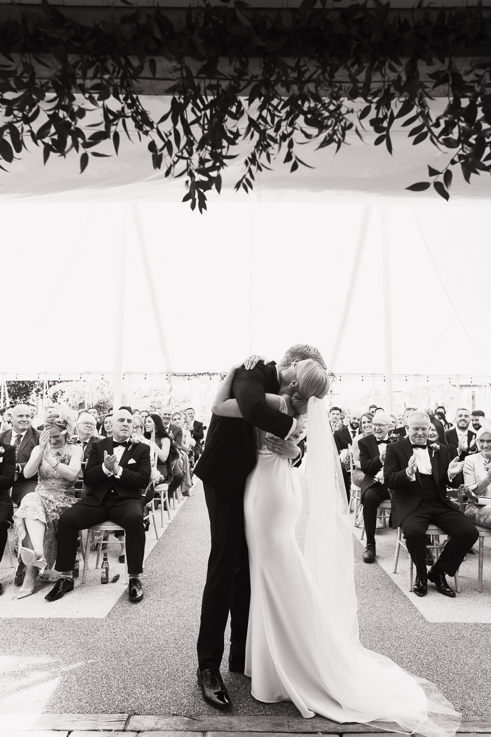 Couple hug at the end of their wedding ceremony. Friends and family clap. Outdoors in the marquee. Suit by The Groom’s Room in Colchester. Dress called Jack by Made With Love from Halo + Wren bridal. Houchins wedding photography by Tracy Morter.