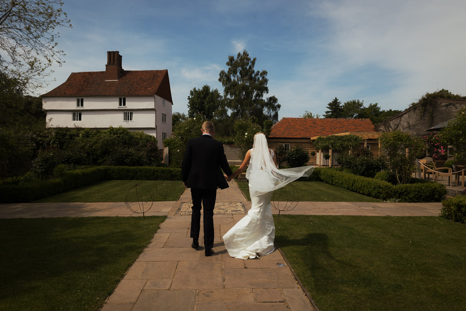Photograph of couple straight after wedding ceremony at an outdoor wedding venue in Essex. A sunny day with blue skies. The bride is wearing a dress called Jack from Made With Love, from Halo & Wren. The groom is wearing a suit from The Grooms Room. Houchins wedding photography in the summer.