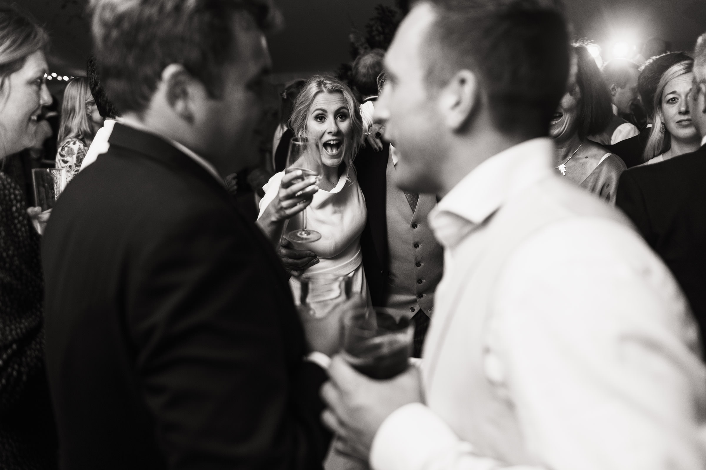 Bride holding a glass of wine surrounded by people on the dance floor. She is wearing a boat neck wedding gown by Jesus Peiro from Miss Bush. A wedding in a marquee from Sail & Peg, on a vineyard overlooking South Woodham Ferrers by the River Crouch. Essex wedding photographer Tracy Morter.