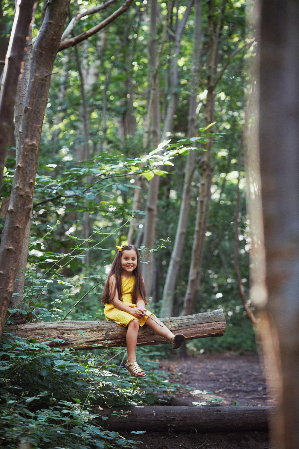 A girl in a yellow dress sitting on a log. She is smiling at the camera. She is surrounded by trees. A natural family photoshoot in Essex. Norsey Woods in Billericay.
