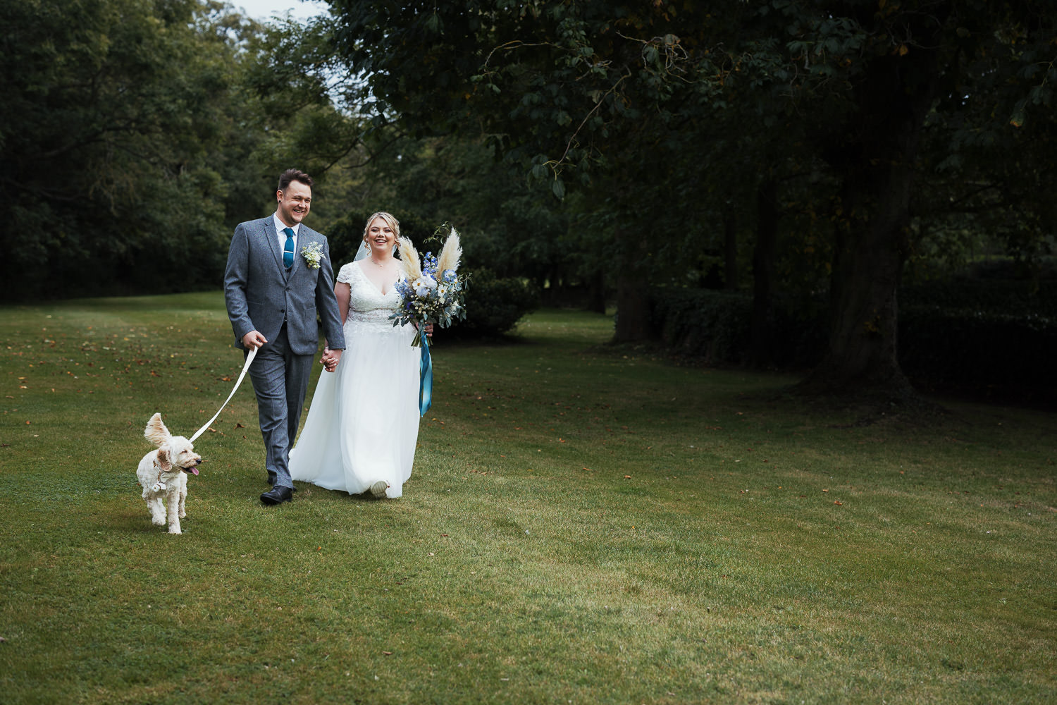 Bride and groom walking in the grounds of Leez Priory with their poodle mix dog. Bride is wearing a dress from Grace & Lace in Chelmsford.