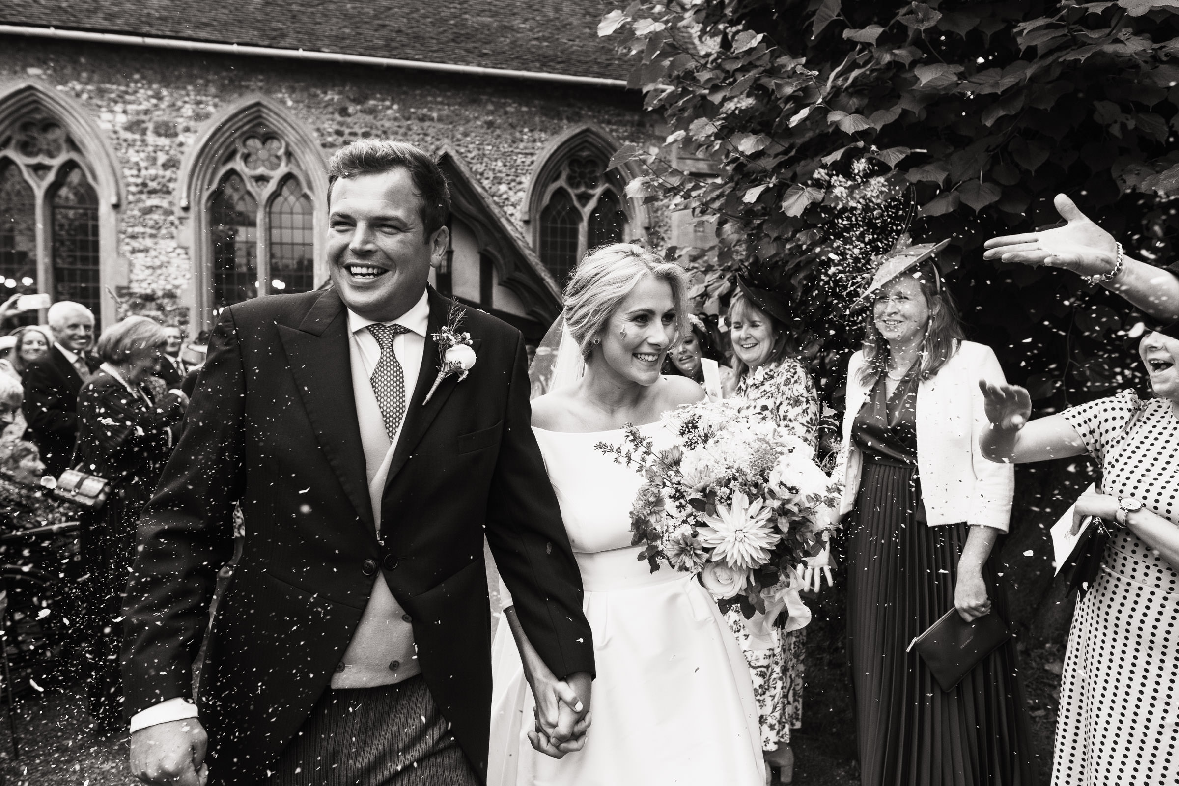 The Parish Church of St John Baptist in Danbury, after a wedding. 

Black and white wedding photography in  Essex of a couple leaving the church. People are throwing confetti.
Bride is wearing a dress from Miss Bush.