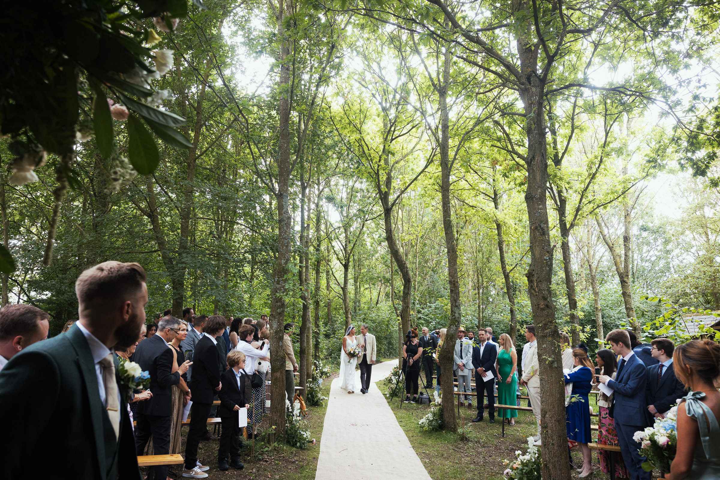 Bride walking down the aisle with her father before a woodland ceremony outside at Alpheton Hall Barns in Suffolk. Surrounded by trees. Bride is wearing Maggie Sottero-Baxley 22MW548B01.