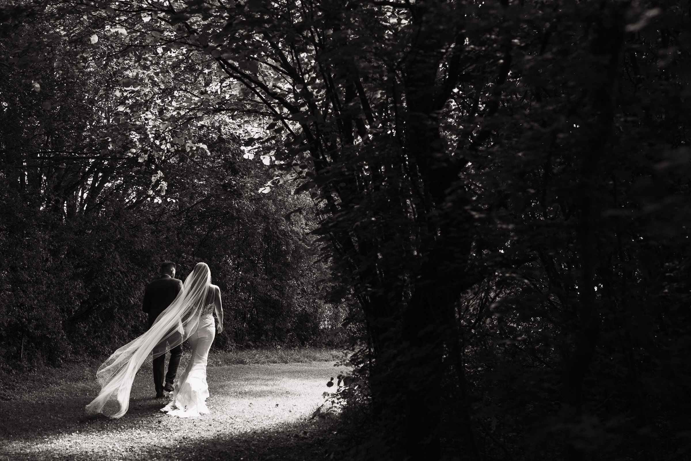 Bride and groom walking down the tree lined lane after a woodland ceremony outside at Alpheton Hall Barns. Surrounded by trees. Bride is wearing Maggie Sottero-Baxley 22MW548B01.