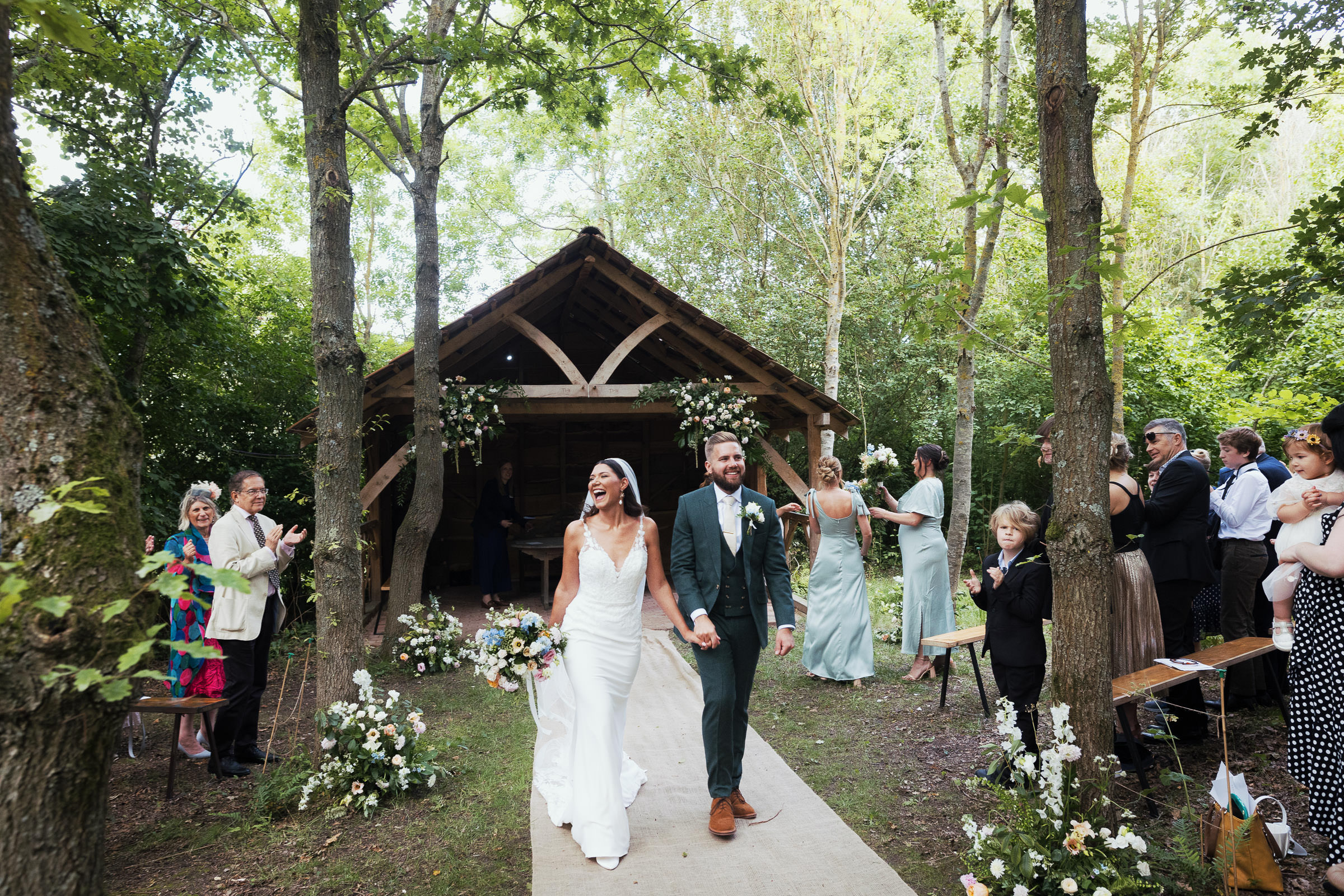 Bride and groom smiling as they walk down the aisle after a woodland ceremony outside at Alpheton Hall Barns. Surrounded by trees. Bride is wearing Maggie Sottero-Baxley 22MW548B01.