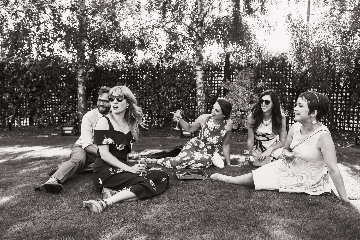 Five wedding guests sat on the grass on a warm day at Crondon Park venue near Chelmsford. Woman with long hair is wearing Warehouse, BUTTERCUP COLD SHOULDER DRESS. Woman in the middle is pointing, wearing a floral dress. Person on right with shorter hair has a pale dress.