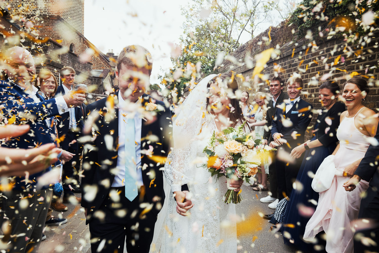 A bride and groom make their way through a confetti tunnel. The confetti is obscuring their faces. After their wedding at St Peter's Church off Church Lane, Petersham Rd, Richmond TW10 7AB.