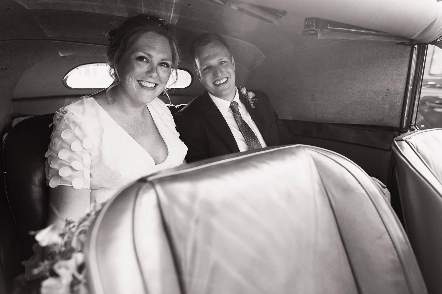 A bride and groom smile at the camera in the back of a car.
After their wedding at the church in Thaxted.
Vagabond wedding dress called Anouk, Circle motif chiffon A line gown with deep V neckline with buttons in the front.