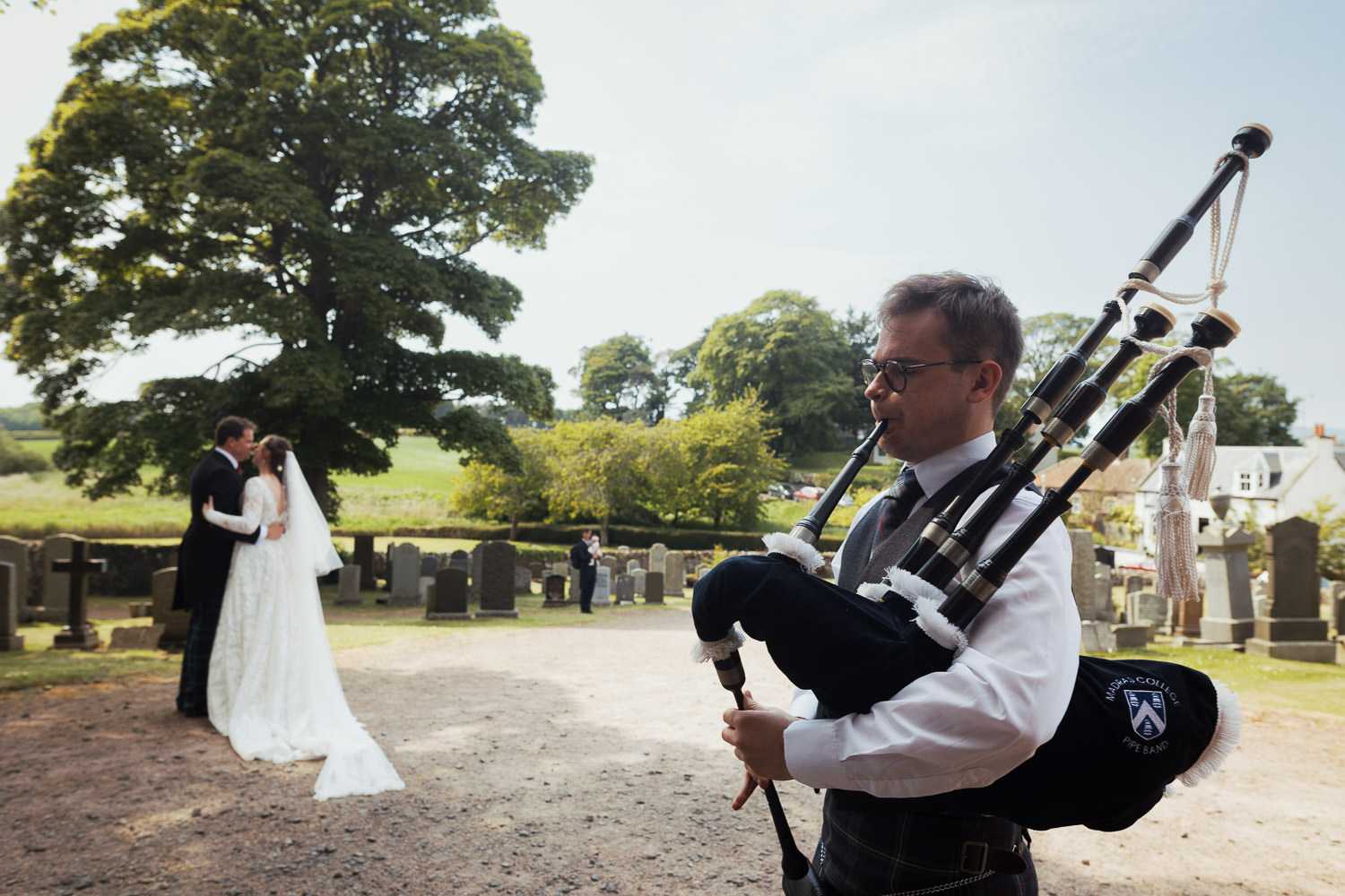 Piper with bagpipes displaying Madras College Pipe Band. Playing as the newly married couple at East Neuk Trinity Church of Scotland Kilconquhar Parish Church kiss near a tree.