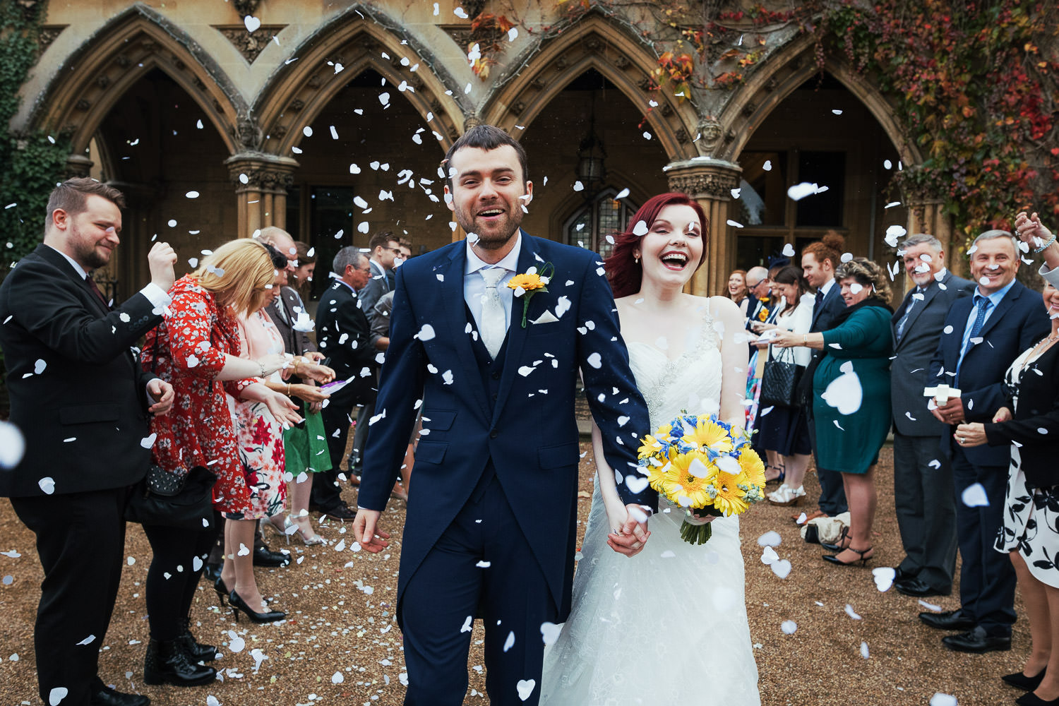 A bride and groom walking through a confetti tunnel. The groom is in a blue morning suit and the bride has red hair. At the wedding venue Manor by the Lake.