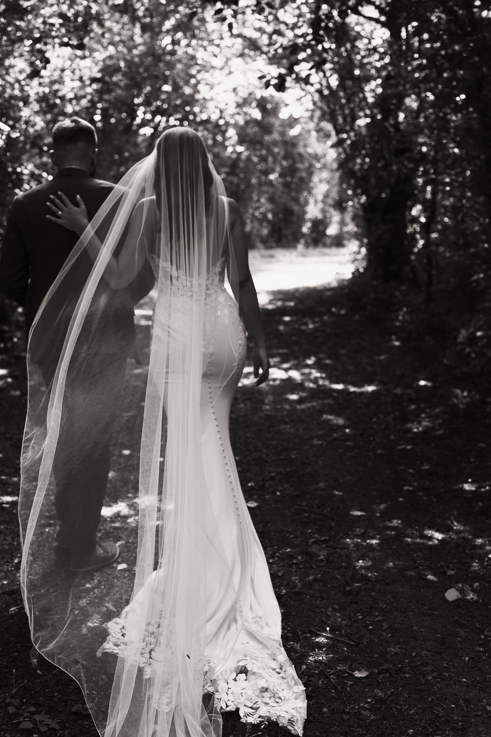 Bride walking with husband, seen from behind wearing Maggie Sottero-Baxley 22MW548B01 backless crepe bridal gown.