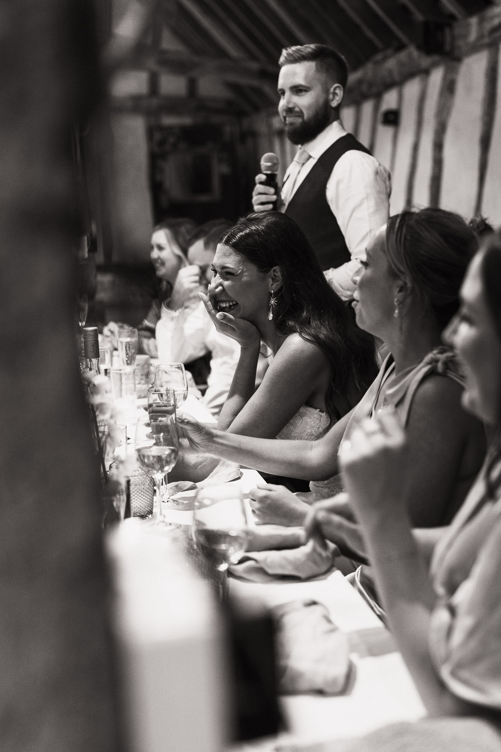 A long top table at Alpheton Hall Barns. Guests and bride are seated as the groom stands for the speech.