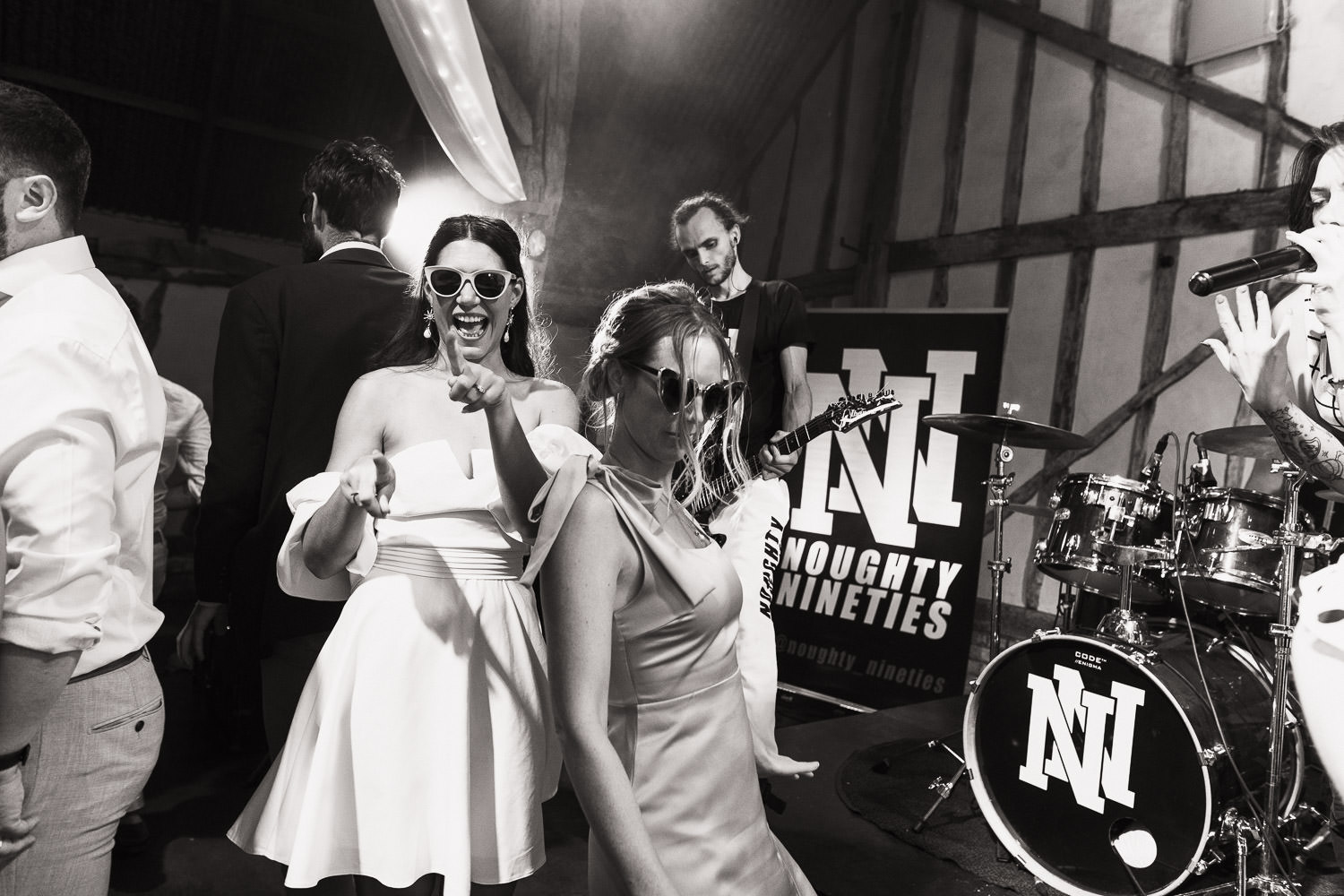 A bride at her wedding reception wearing ASOS Trendyol bandeau sleeve mini dress in white and sunglasses. With her bridesmaid. They are dancing to the band Nought Nineties at Alpheton Hall Barns. Taken by Essex wedding photographer.