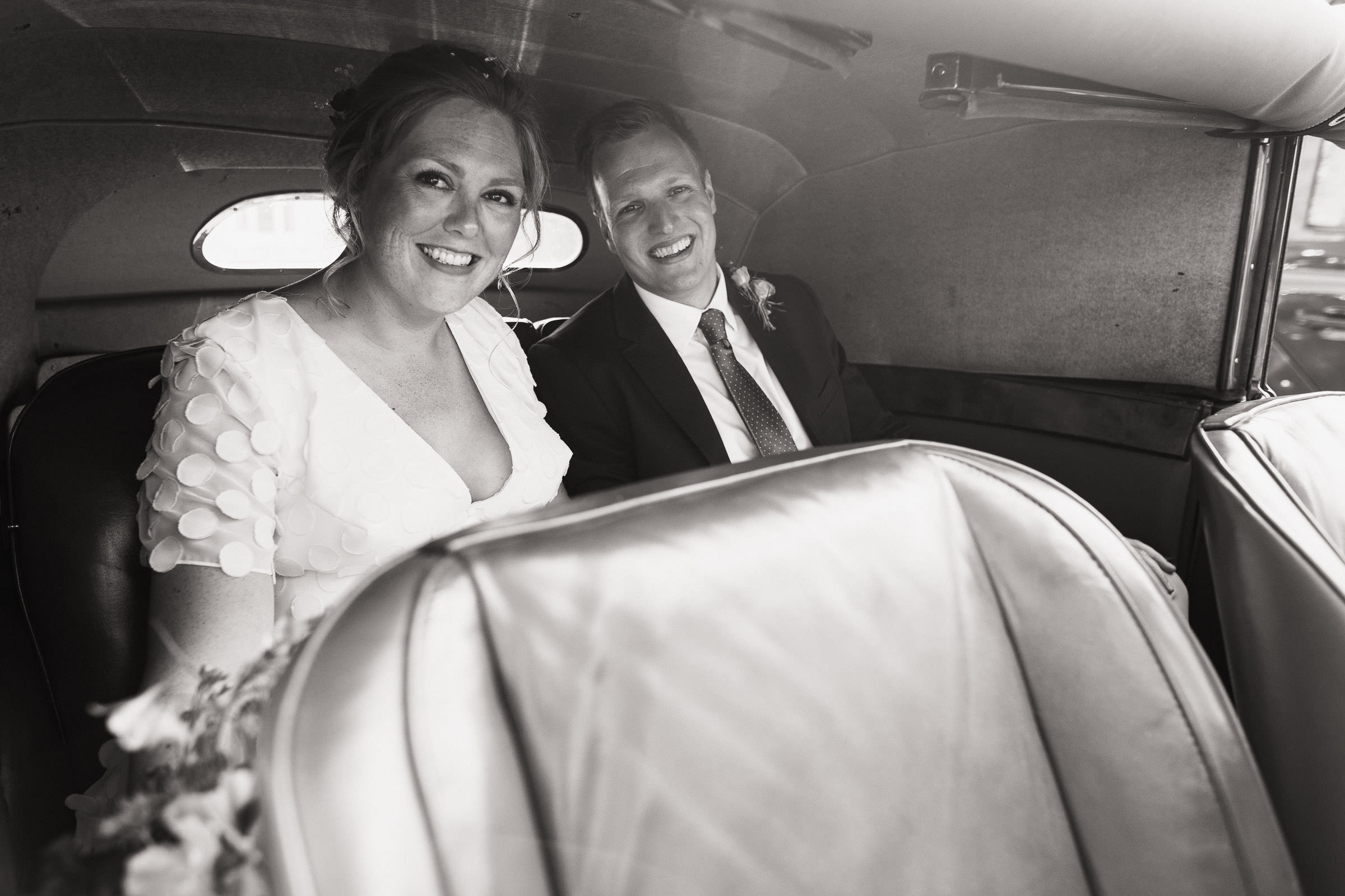 A bride and groom smile at the camera in the back of a car, 1951 Jaguar. After their wedding at the church in Thaxted in Essex. Vagabond wedding dress called Anouk, Circle motif chiffon A line gown with deep V neckline with buttons in the front.