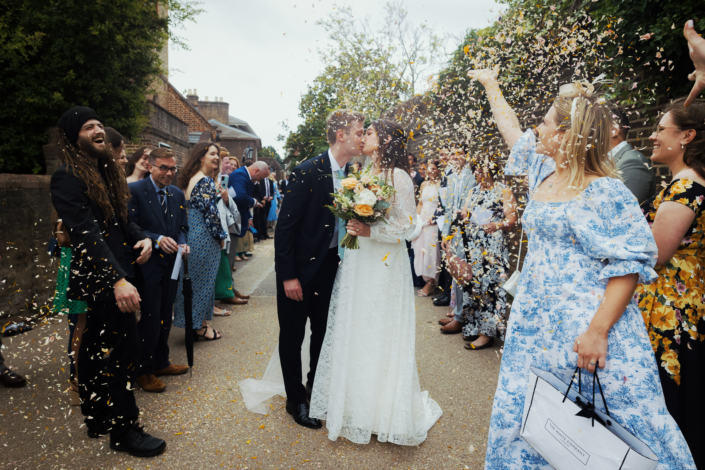A woman with blonde hair wearing a Nothing Fits But Kiko Dress is throwing confetti over the kissing newly married couple. A wedding at St Peter's Church in Petersham. On the left a woman is wearing the Rixo midi dress called Jeanie.