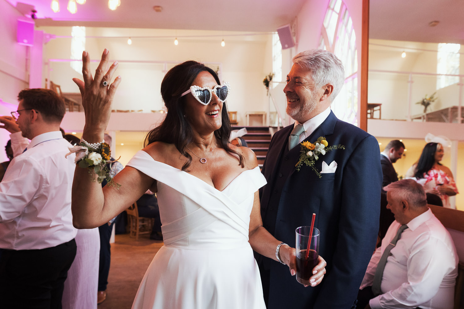 New wife wearing off-the-shoulder Ivory dress with pockets by Justin Alexander called Denley. Sunglasses and holding a drink, holding other hand in the air as her husband in a navy suit smiles. The Old Parish Rooms wedding photography.