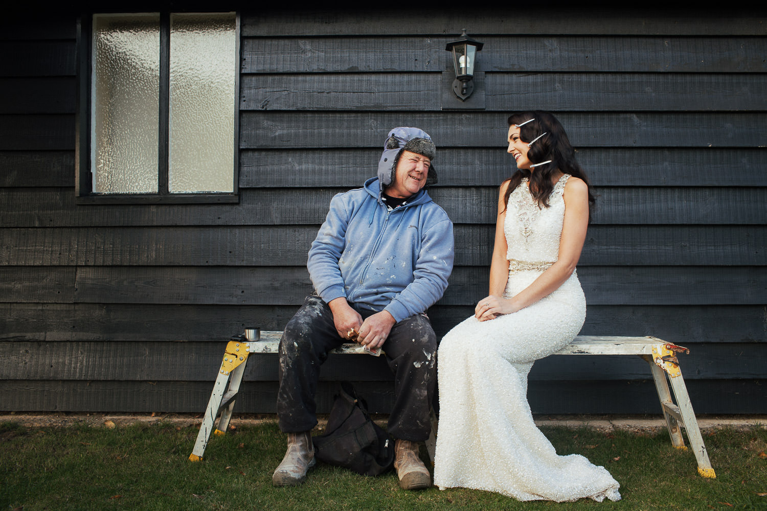 At the wedding venue High House near South Woodham, Amelia in a wedding dress sits on a workbench with a handyman on his break.