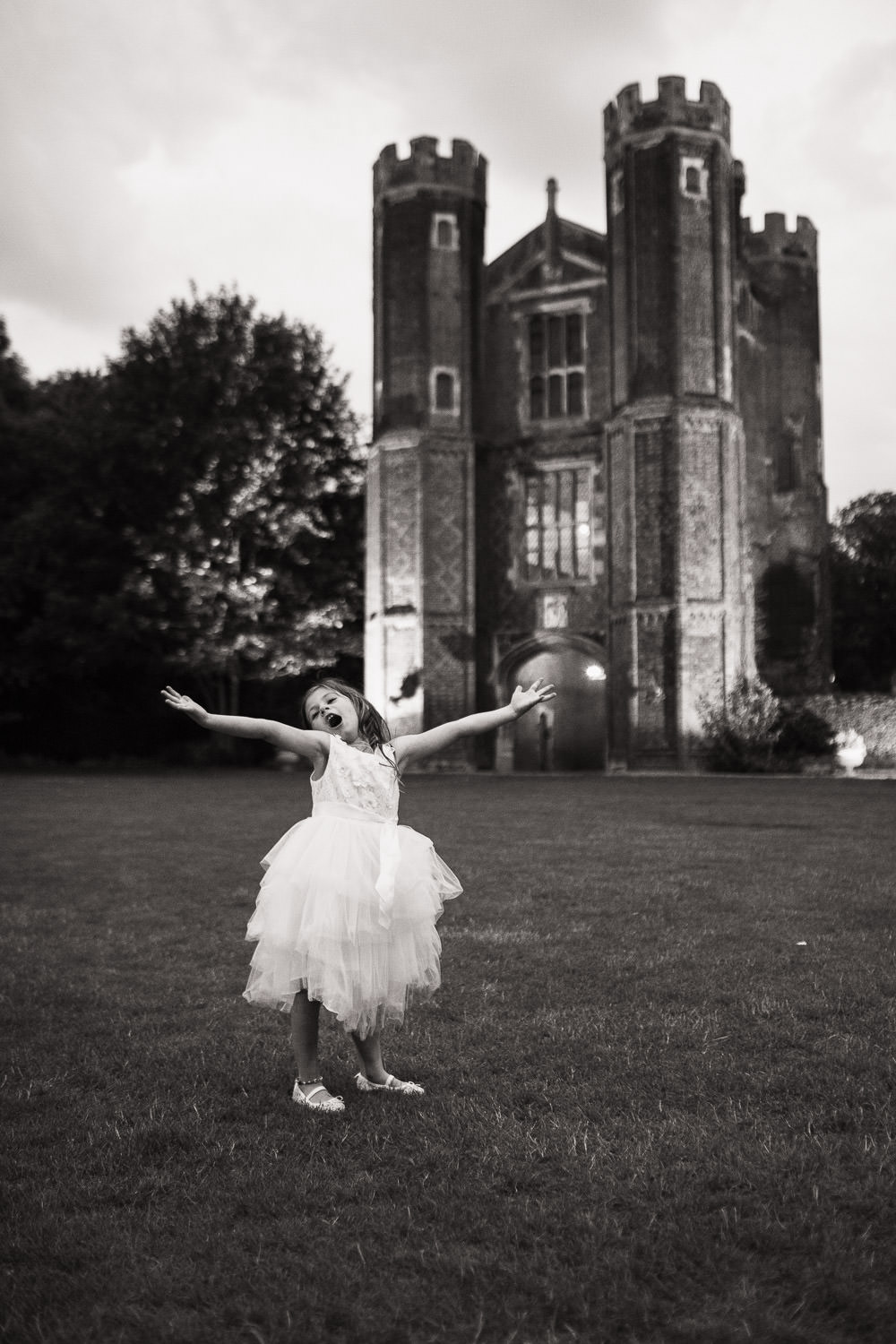 A flower girl in a white dress with her arms out, singing. The Great Tower at Leez Priory behind her.