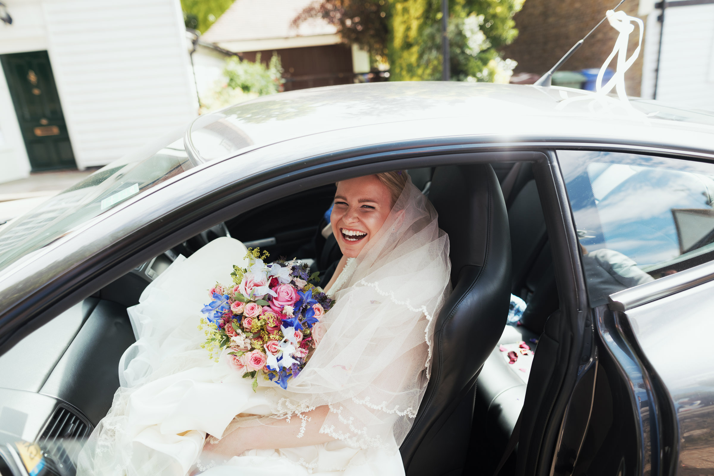 Bride getting in the car smiling after her wedding at St Giles & All Saints Church, Orsett. Holding a bouquet, wearing the Morilee Laurie ML5684 wedding dress, and a veil.