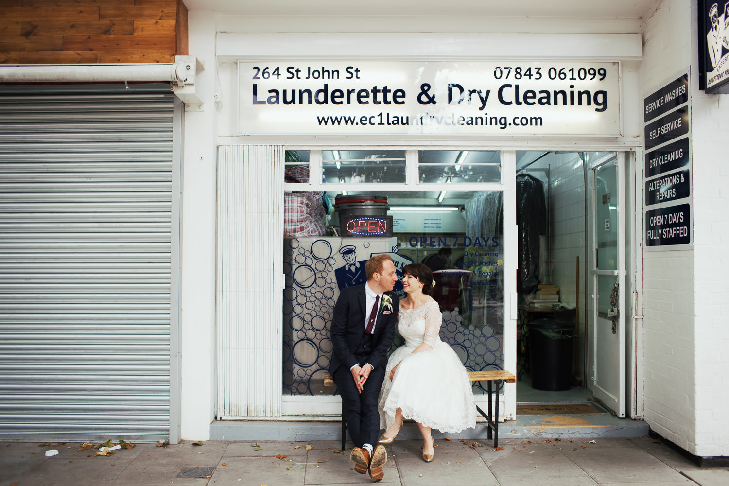 Bride and groom sat on a bench outside 264 St John Street Launderette & Dry Cleaning.