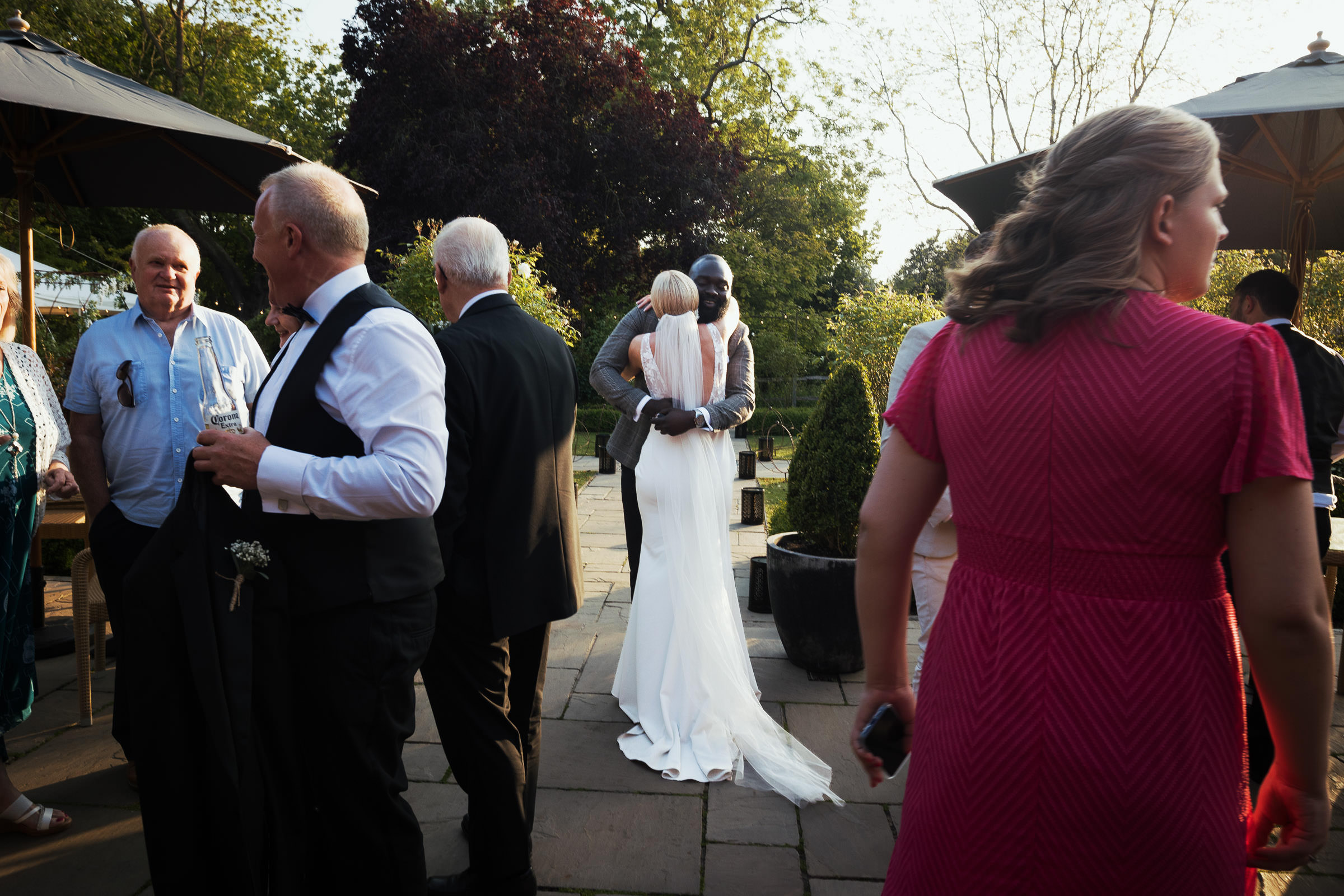 Documentary wedding photography by Tracy Morter at the Essex wedding venue Houchins. The bride is wearing a dress called Jack by Made With Love Bridal from the Halo + Wren bridal shop. Bride is hugging a wedding guest on the terrace in the evening.