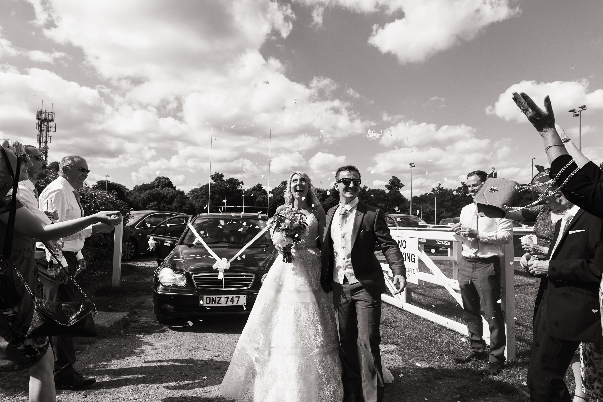 Newly married couple arriving at The Cobham Curve for their wedding reception. Their guests throwing confetti over them as they walk past. It's a sunny day with blue skies. Woman wears Allure Bridals ball gown 9400.