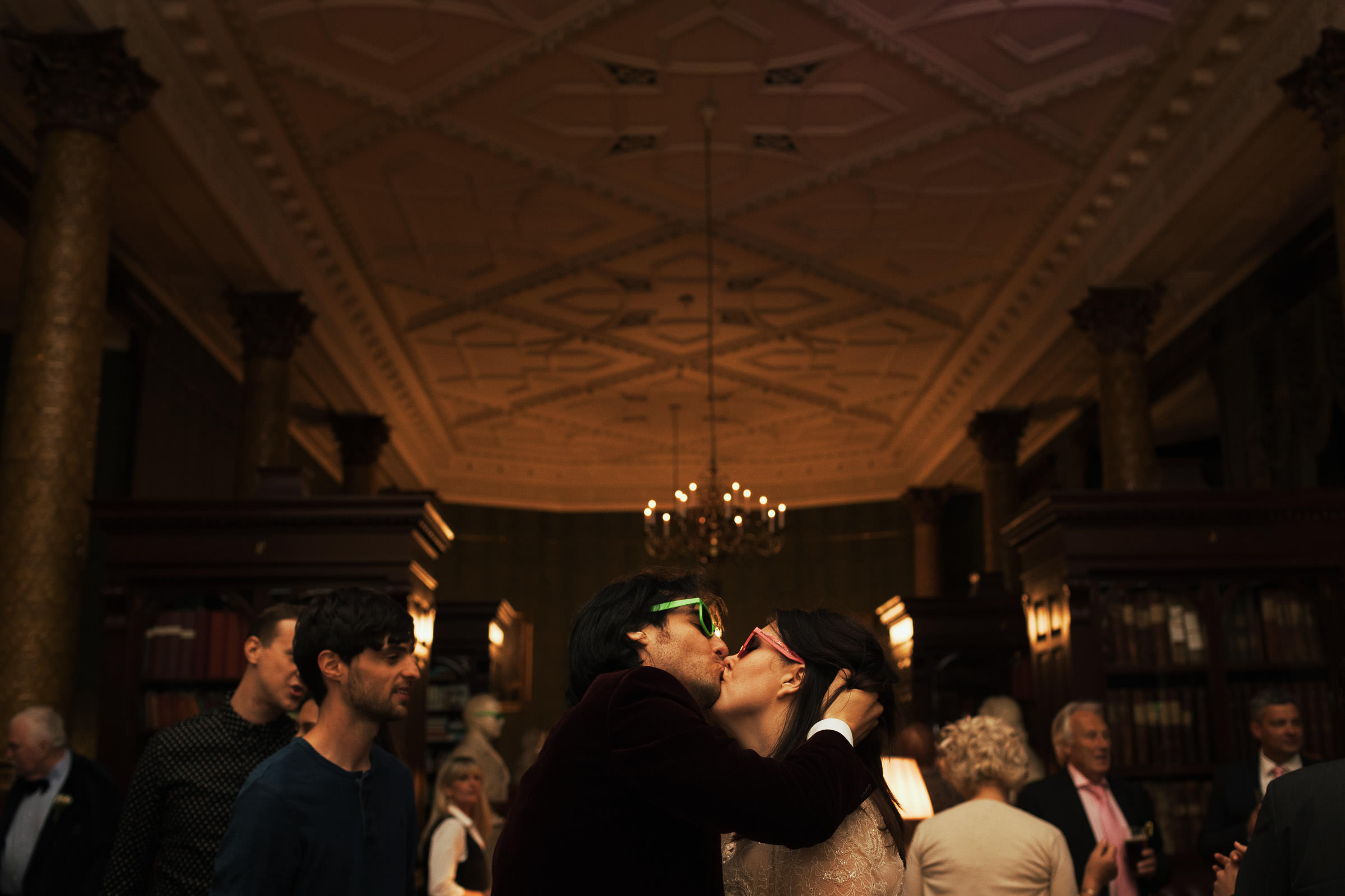 Newly married couple wearing bright sunglasses at wedding reception, kissing on the dance floor of the smoking room of the National Liberal Club.