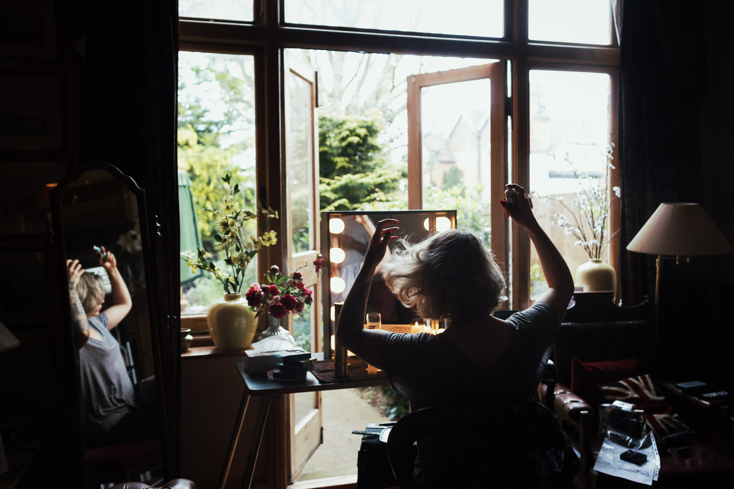 A blonde woman sitting at a dressing table spraying her hair, getting ready for a wedding. The doors to the room are open to the garden. London wedding photography.