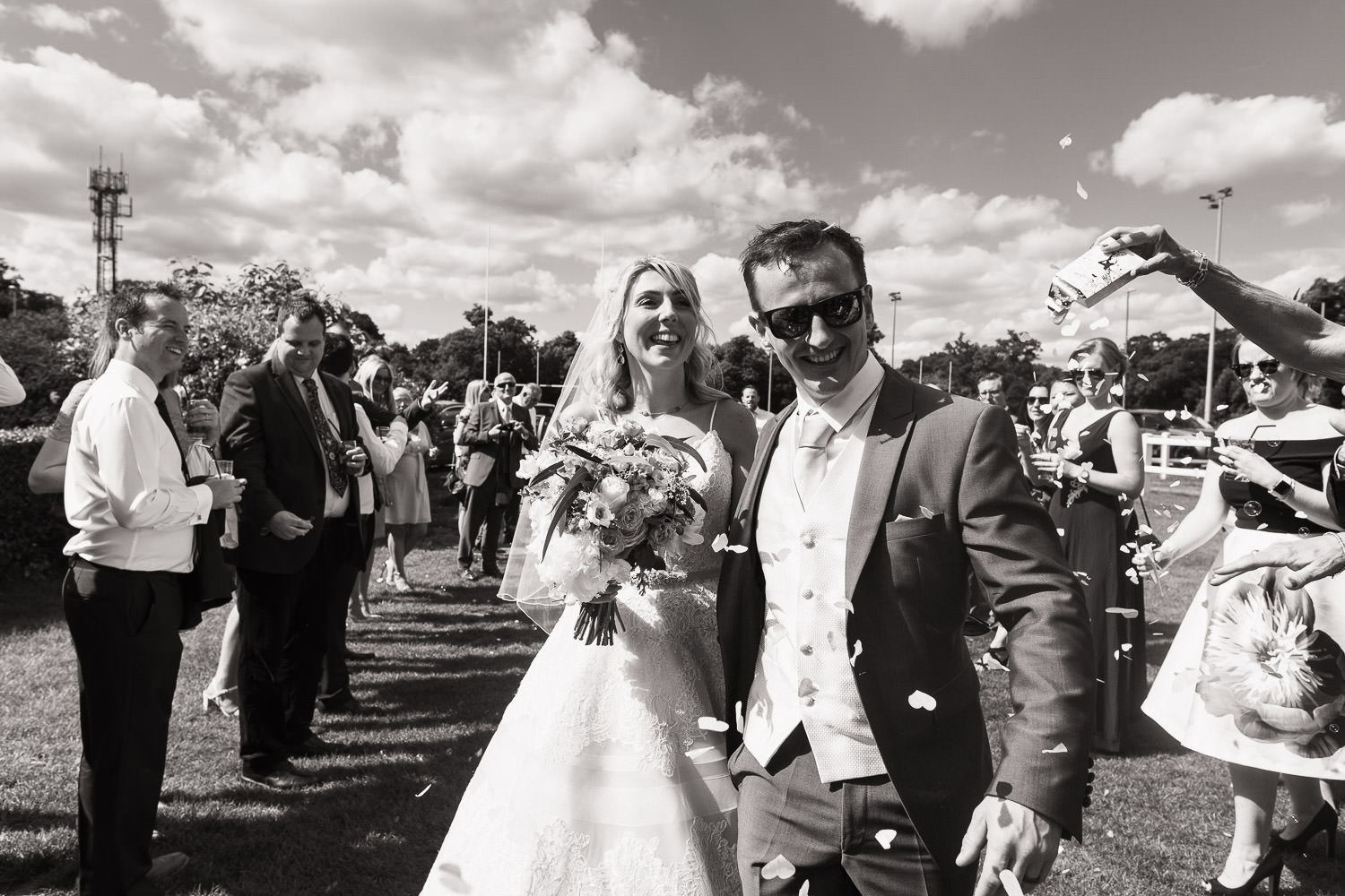 Newly married couple arriving at The Cobham Curve for their wedding reception. Their guests throwing confetti over them as they walk past. It's a sunny day with blue skies. Woman wears Allure Bridals ball gown. Groom is wearing a suit and sunglasses.