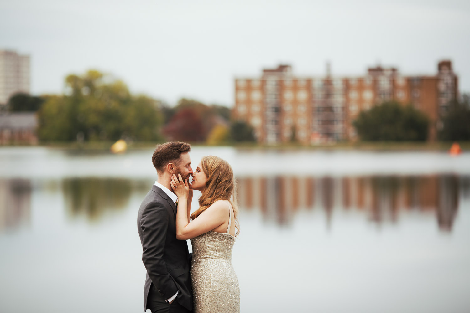 Newly married couple almost kissing by the lake at the West Reservoir Centre in Stoke Newington. the view of buildings in the Woodberry Down area. Bride has long hair and is wearing a sparkly dress. Man has hands in pockets of his suit.
