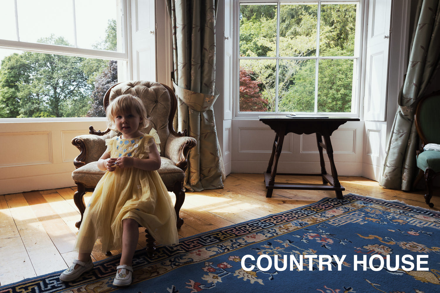 Documentary style wedding photograph at a country house wedding.

A little girl in a yellow dress is sitting on a stool in the bedroom of a wedding venue called Homme House. There are windows and trees in the background.JoJo Maman Bébé Floral Yellow Floral Tulle Party Dress.