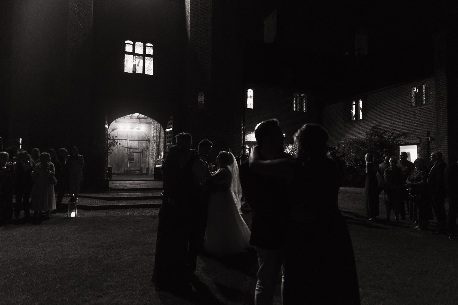 In the evening a bride and groom are having their first dance outside on the lawn. Their friends and family have joined in with the slow dance. They are illuminated by the light from the Leez Priory carriageway.