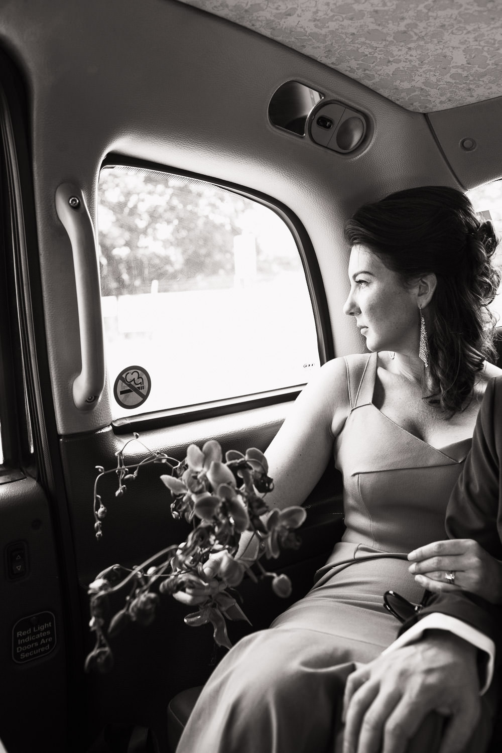 A bride in the Roland Mouret Mermaid Dress in a taxi. Holding orchids and looking out of the window. Her arm holding the elbow of her husband out of the shot. On their way to their Petersham Nurseries wedding breakfast. Documentary wedding photo taken in London.
