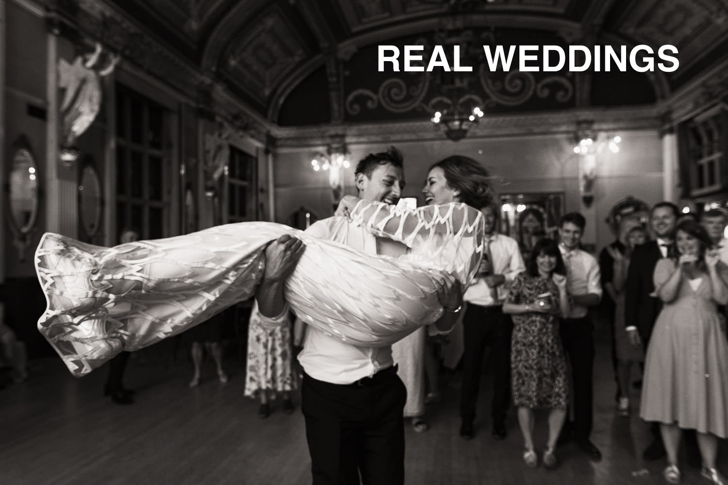 REAL WEDDINGS title photo. Groom picks up his wife and spins her around at a wedding reception at the Old Finsbury Town Hall. The bride, wearing the Willow dress from Halfpenny London. Captured in a documentary wedding photography style.