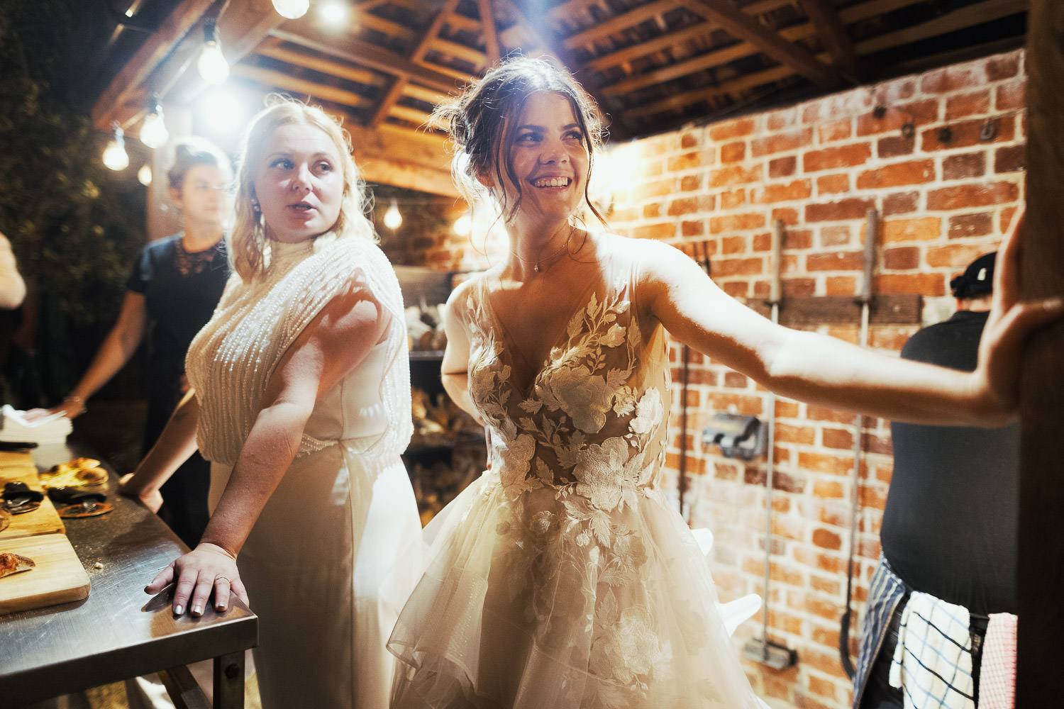Brides stand by the illuminated pizza oven in the courtyard at Alpheton Hall Barns during the evening of their wedding day. The bride in the foreground is wearing a WTOO by Watters Montgomery dress (STYLE #12716).