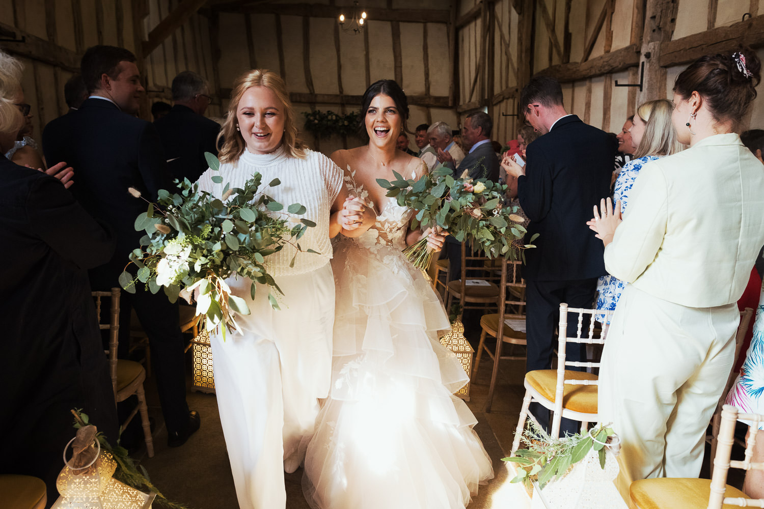 Joyful newlyweds walk down the aisle following their Suffolk Registrar wedding ceremony at Alpheton Hall Barns. One bride wears a WTOO by Watters Montgomery dress (Style #12716), while the other is adorned in a Joanna Hope Beaded Bridal Jumpsuit. Beautiful flowers by Wild + Mae complement the scene.