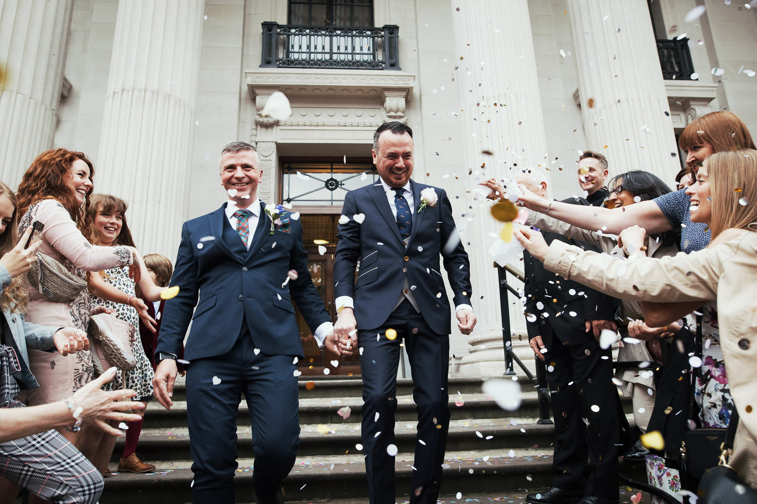 At Old Marylebone Town Hall, newlywed husbands descend the steps holding hands amidst confetti thrown by guests.