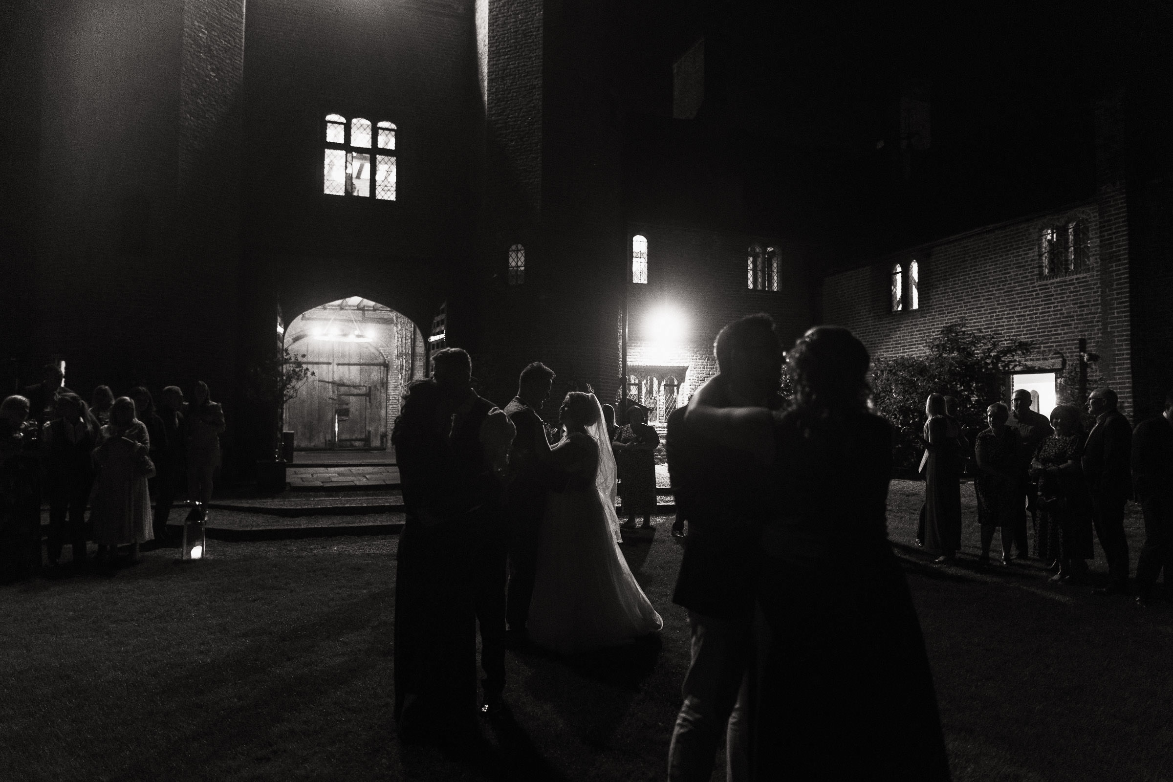 In the evening a bride and groom are having their first dance outside on the lawn at their wedding reception. Their friends and family have joined in with the slow dance. They are illuminated by the light from the Leez Priory carriageway.