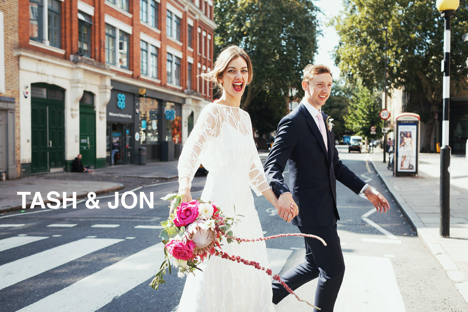 Bride and groom crossing Tysoe Street near Exmouth market. The woman is wearing a Halfpenny dress and is holding pink flowers, she is smiling widely at the camera. The groom is wearing a dark suit and pale pink tie. The Co-op and a phone box are in the background. Wedding photojournalism style photograph in London.