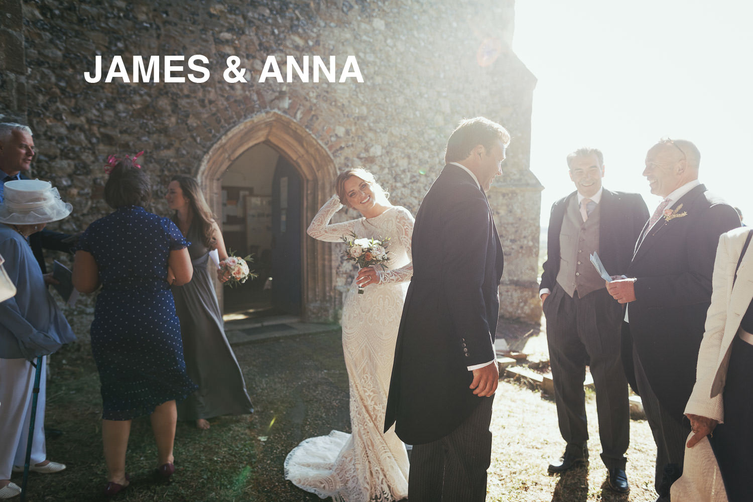 Wedding at The Church of St Mary & St Margaret in Stow Maries, near South Woodham Ferrers. Guests chat while bride, illuminated by sunlight, looks at camera. She wears a bespoke long lace wedding dress with sleeves by Louise Sullivan (Louise Bridal) dressmaker.