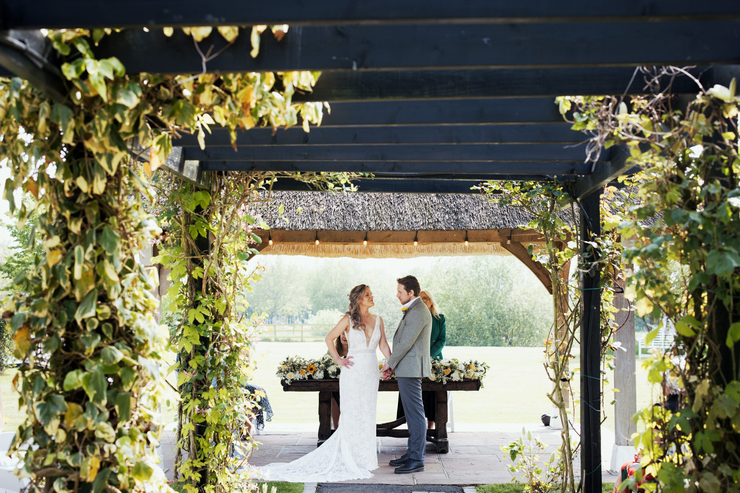 Bride and groom holding hands during their wedding ceremony outdoors under the thatched gazebo. Essex registrar in the background. Green foliage decorating the pergola framing them. Bride wearing a lace dress by Demetrios J’Adore Bridal Wear. Groom wearing Moss herringbone tweed waistcoat and jacket in sage. Samantha May Floristry. High House Weddings, Old Heath Rd, Althorne, Chelmsford CM3 6EW. Now with new Farmhouse Accommodation in 2024 and an orangery in 2025.