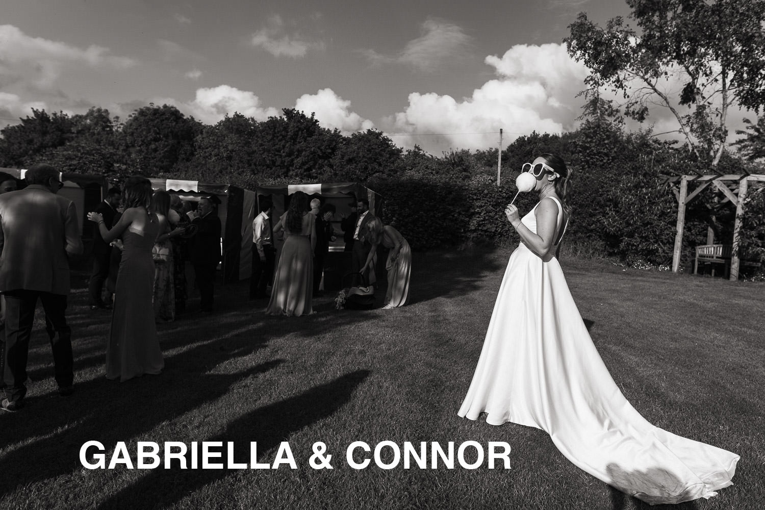 In her white satin wedding dress and oversized sunglasses a bride eats candy floss in the the sun. Guests play funfair games. At Alpheton Hall Barns wedding venue.