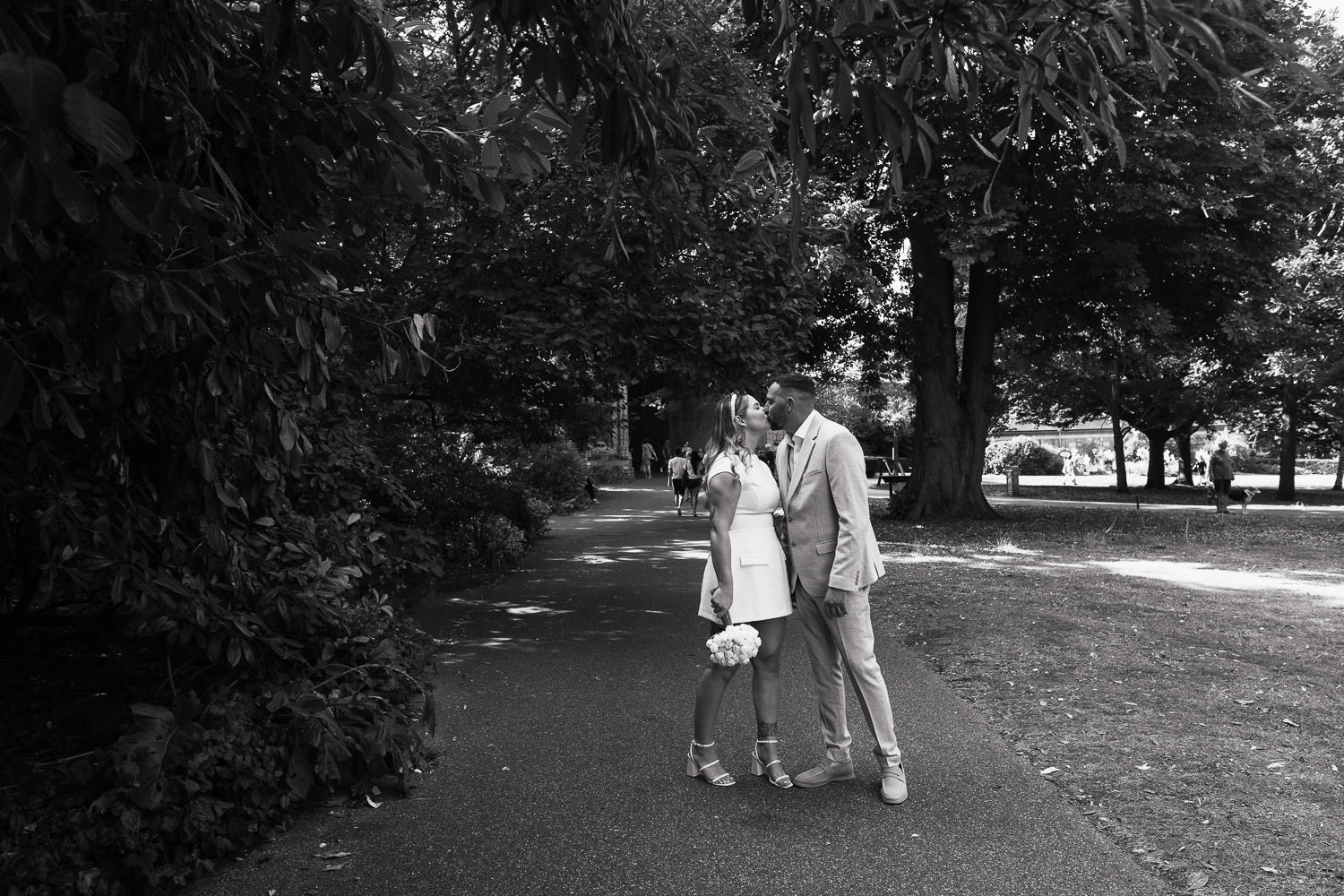 After a Bury St Edmunds Register Office Suffolk wedding ceremony the newly married couple kiss in Abbey Gardens.Bride is wearing the Odd Muse The Ultimate Muse Cap Sleeve Mini Dress in White. Groom is wearing a Moss slim fit off white check suit.