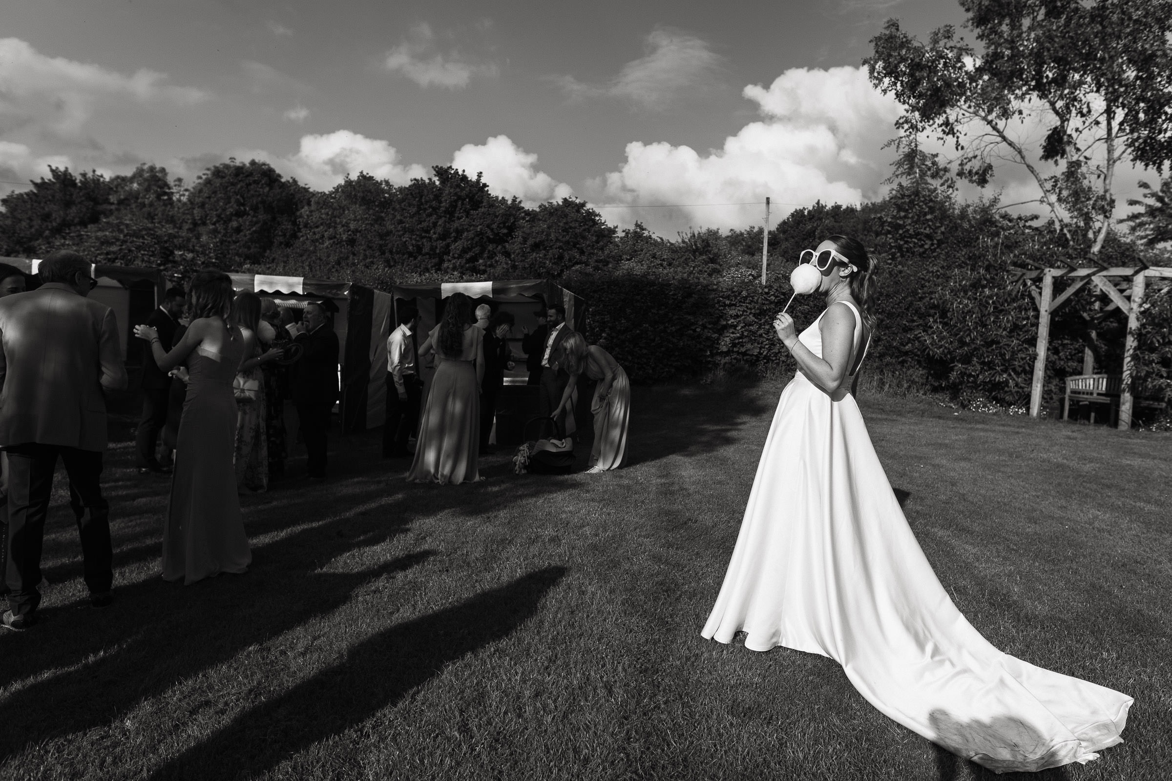 In her white satin wedding dress and oversized sunglasses a bride eats candy floss in the the sun. Guests play funfair games. At Alpheton Hall Barns wedding venue.