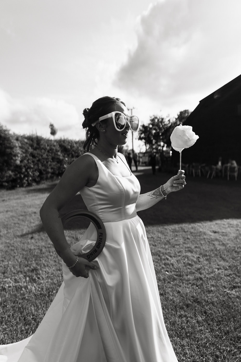 A relaxed bride in oversized sunglasses holds a stick of candy floss and a frisbee, in the bright sunshine outside Alpheton Hall Barns in the stackyard wedding garden. By Essex Wedding Photographer Tracy Morter.