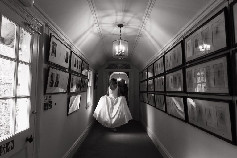 At the Jockey Club Rooms in Newmarket the bride walks down a corridor holding up her wedding dress. Rosa Clará wedding dress with deep-plunge V-neckline and pockets. from BOA Boutique in Surrey. Pictures on the wall, the glass reflecting the bride. Documentary wedding photography near Cambridge.