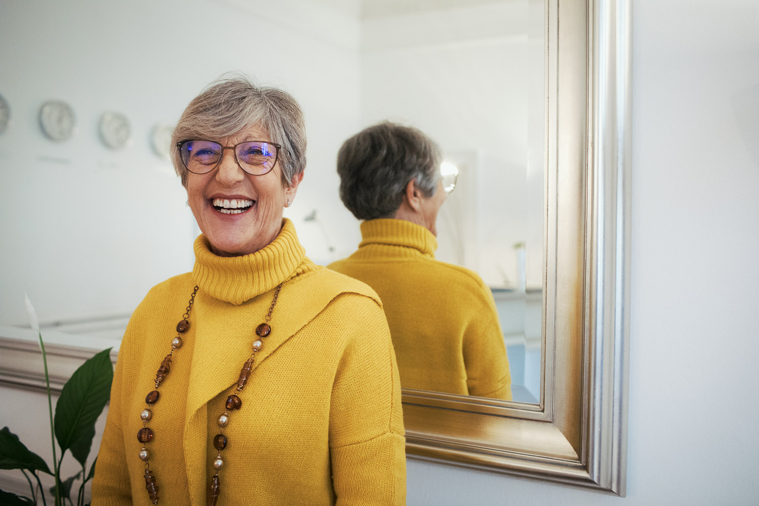 Receptionist wearing a mustard coloured jumper and beaded necklace, at work in the office in front of a mirror. She is laughing.