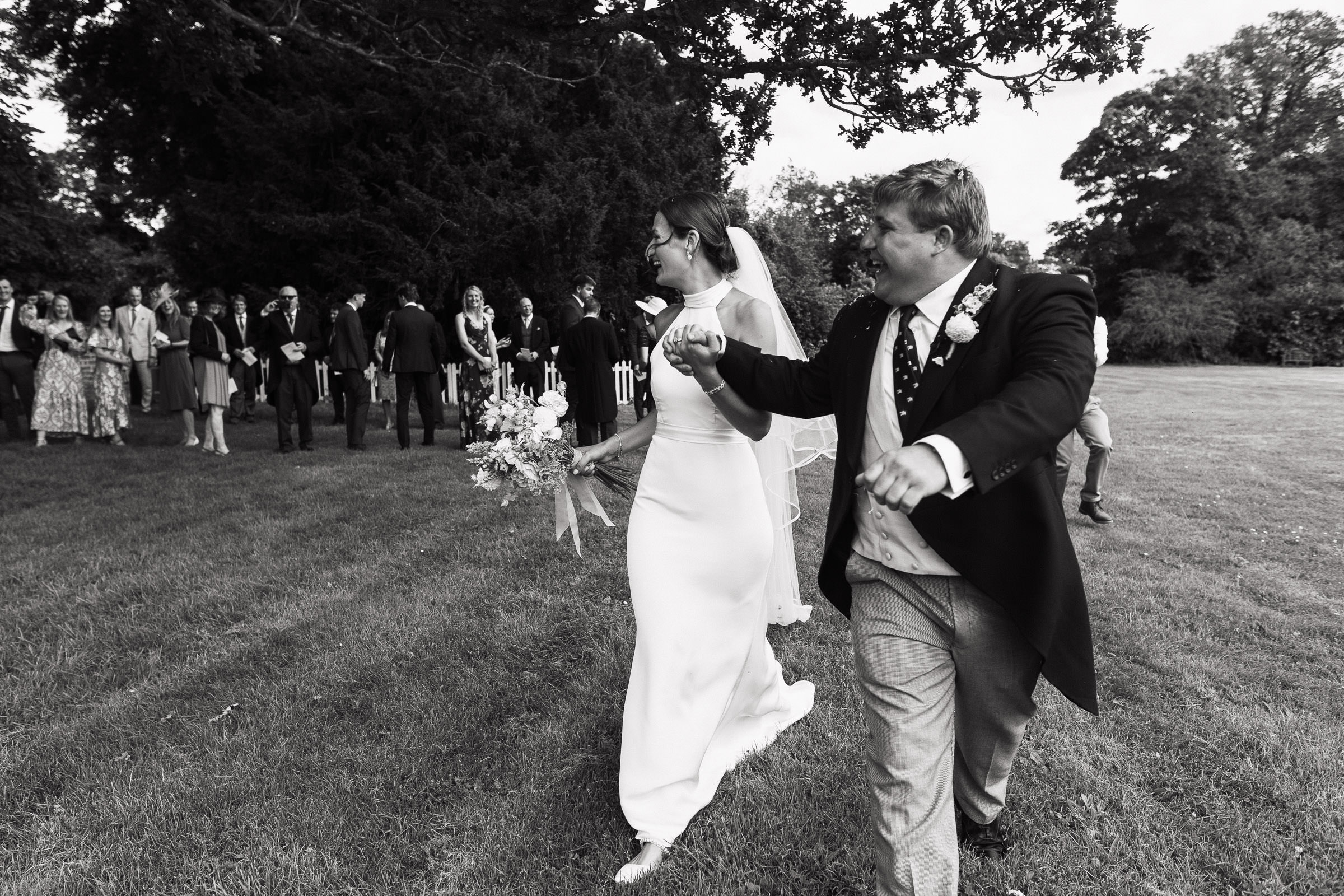 A joyful newly married couple walking away from their guests at the Church of St Peter & St Paul, Heydon. Bride wearing a white halterneck dress by Katrine Mogensen. Documentary wedding photography.